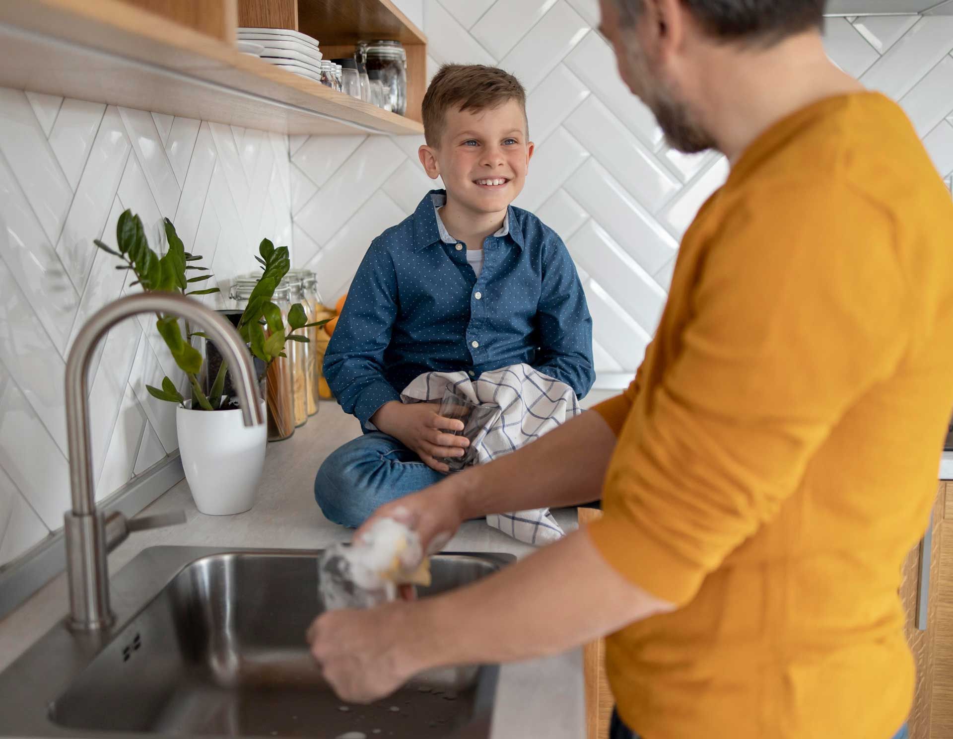 A man washes dishes in the kitchen while a boy sits on the counter, smiling and holding a dish towel.