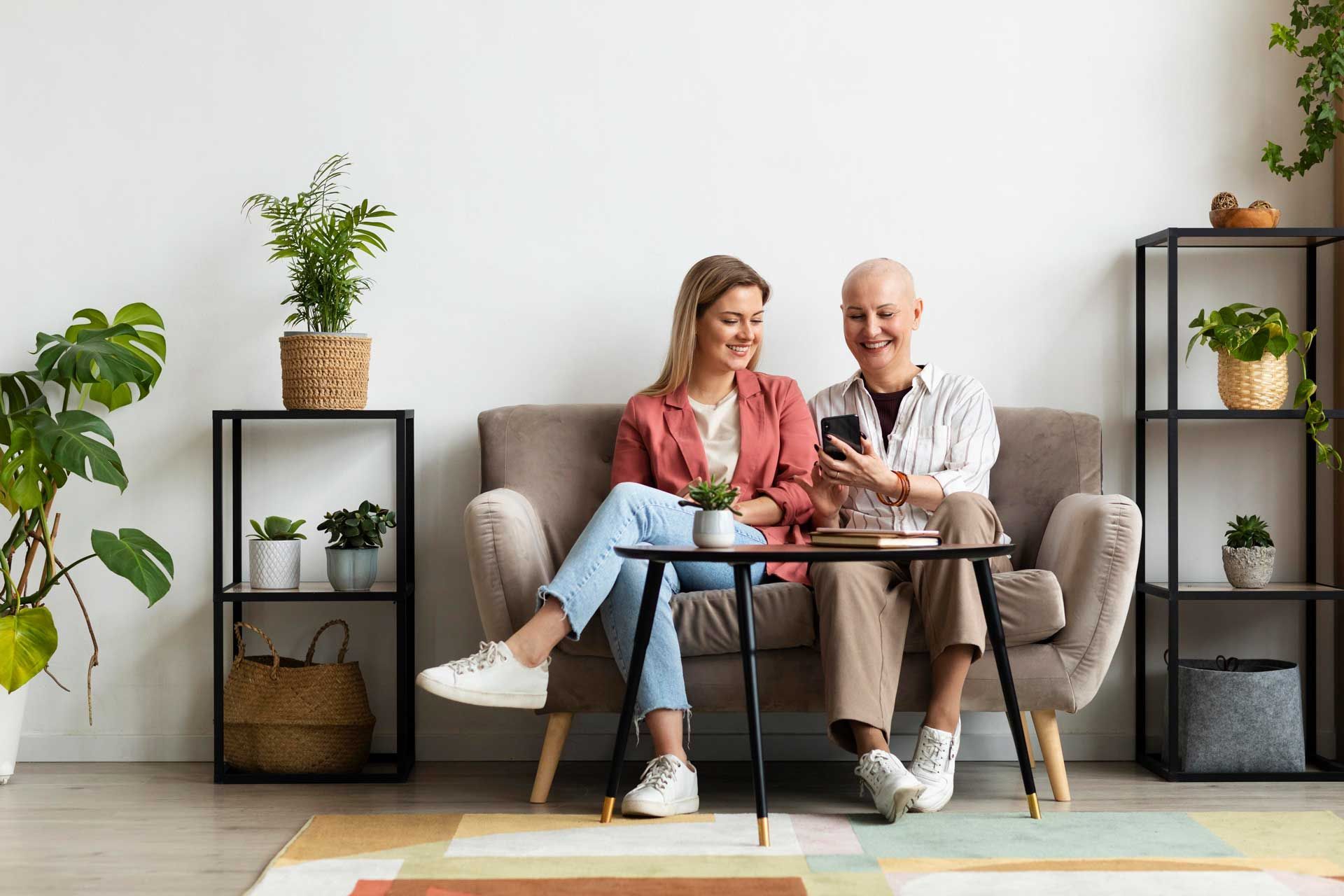 Two women sitting on a couch looking at a phone, laughing. Plants and shelves in the background.