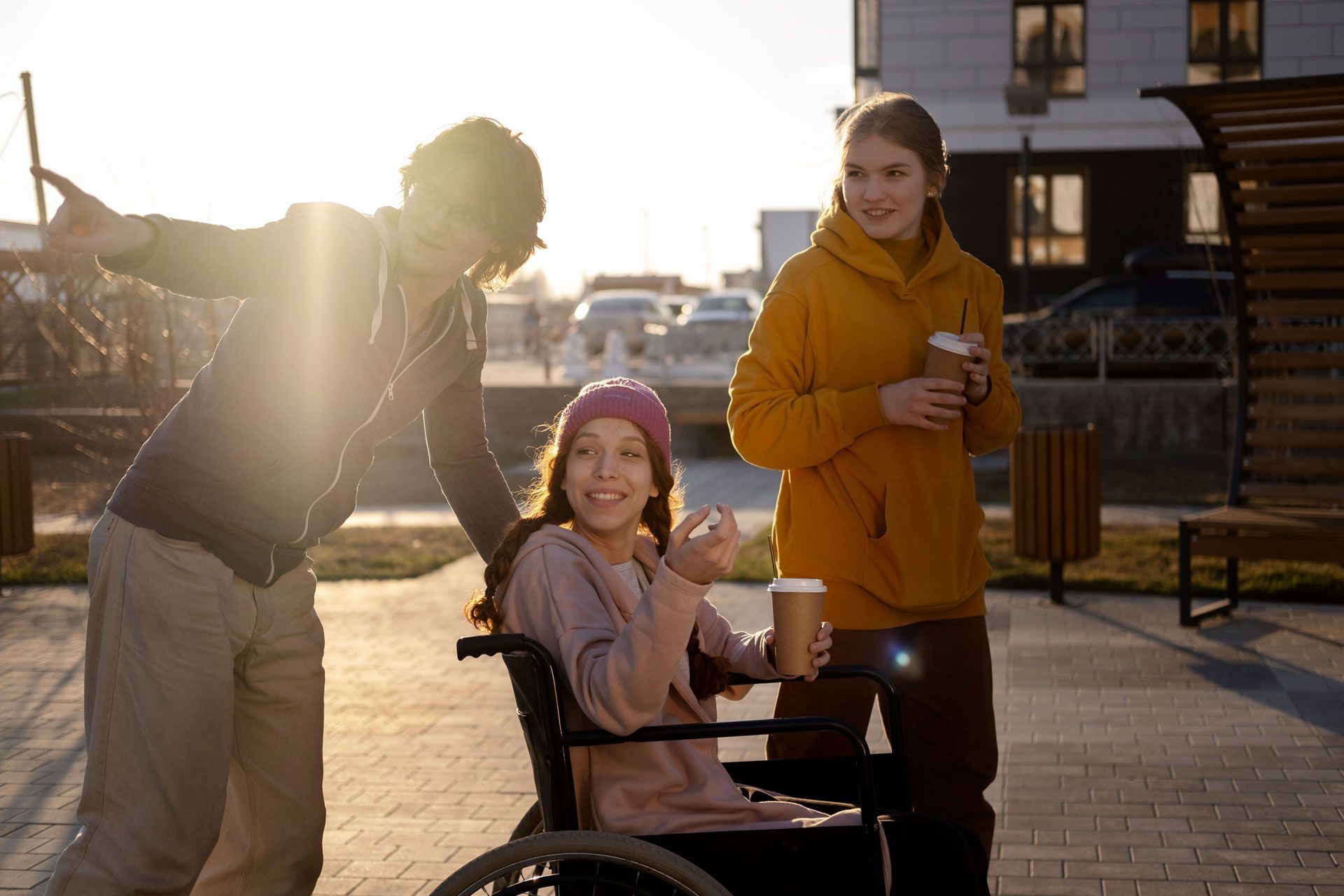 Three people outdoors: One in wheelchair, two standing, with coffee cups, at sunset.