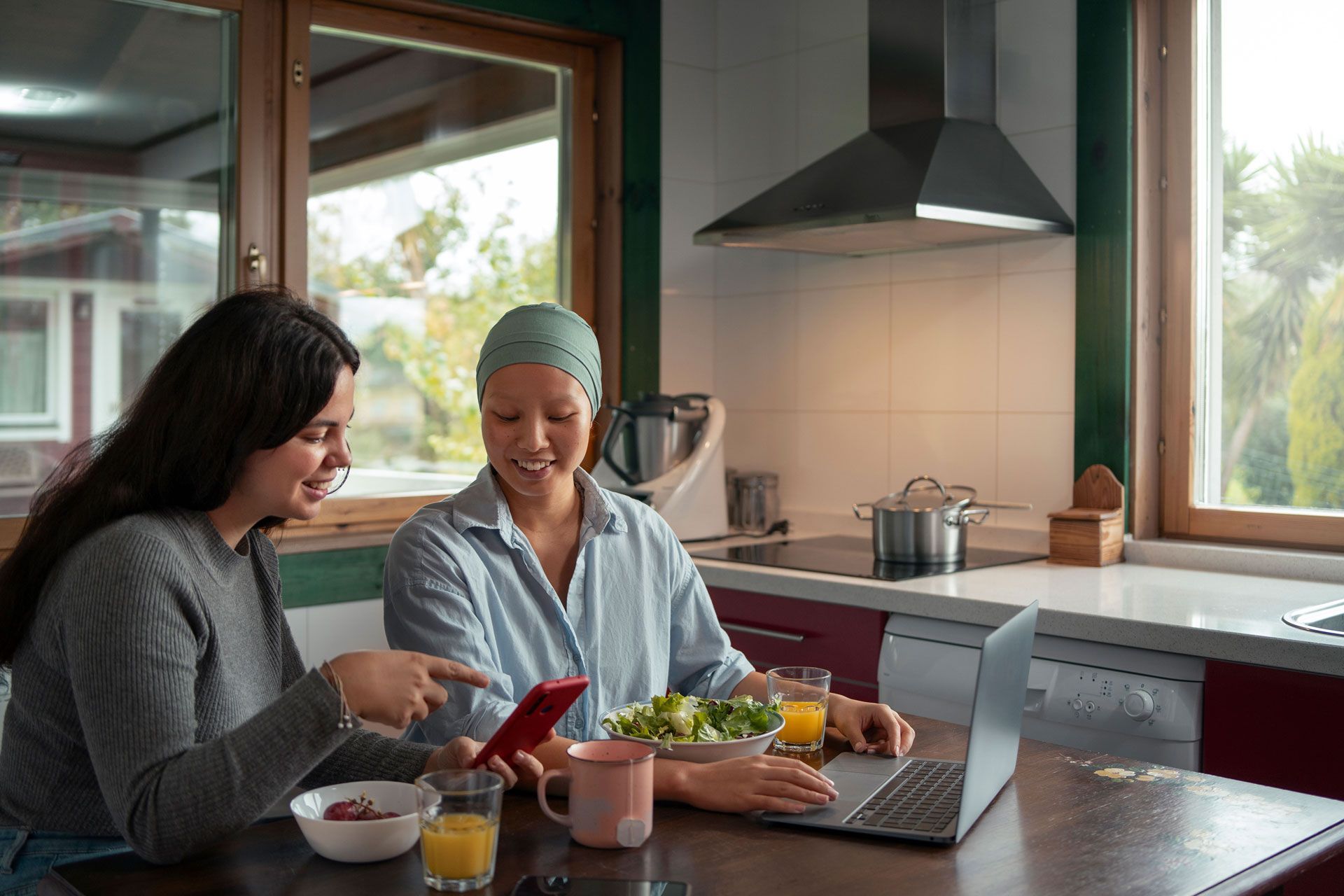 Two women in a kitchen, one pointing at a phone, other using a laptop, both smiling.
