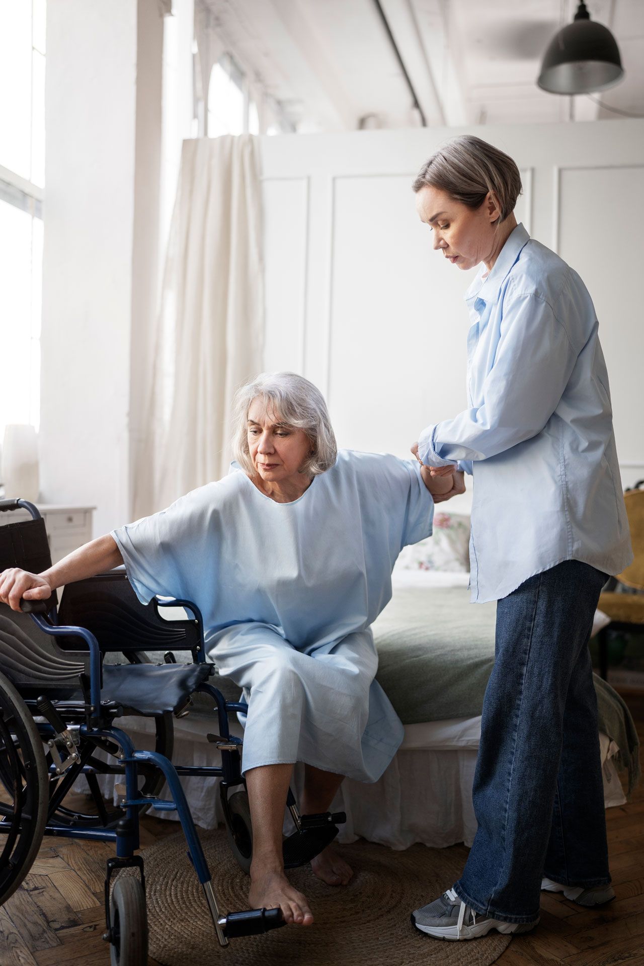 Woman helps an elderly woman from a wheelchair to a bed in a bright room.
