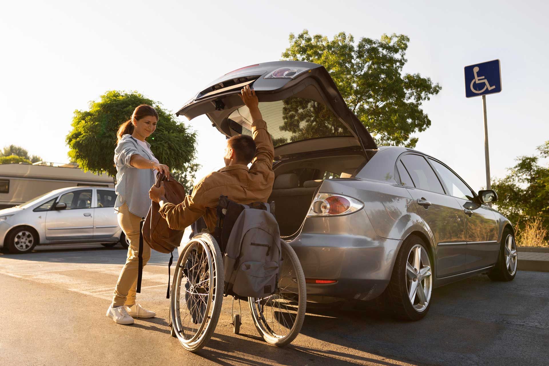 Woman helping a man in a wheelchair load a bag into a car trunk in daylight.