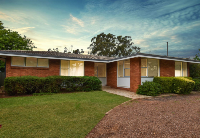 Brick ranch-style house with green lawn and shrubs under a sunset sky. Driveway on the right.