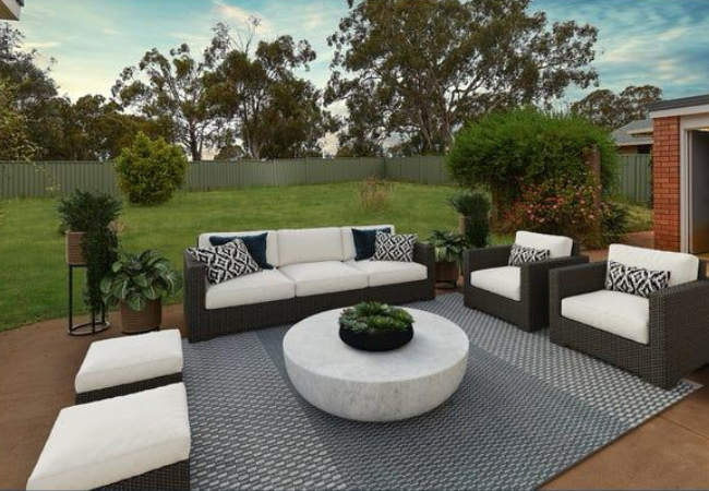 Outdoor patio with a sofa, armchairs, and a circular coffee table on a rug; lush green lawn in the background.