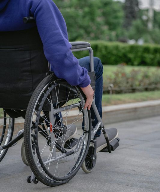 Person in a wheelchair rolling along an outdoor path, wearing a blue sweatshirt and jeans.