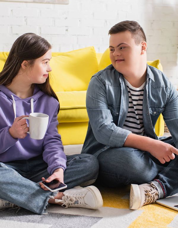 Two young people with Down syndrome sitting on the floor, talking. One holds a mug, the other smiles.