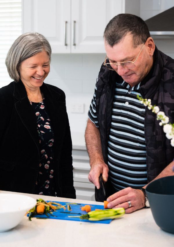 A man and woman in a kitchen, the man slicing vegetables on a cutting board.