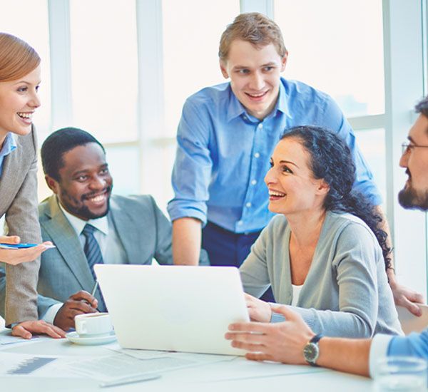 Team of diverse colleagues collaborating around a laptop, smiling and engaged in a bright office.