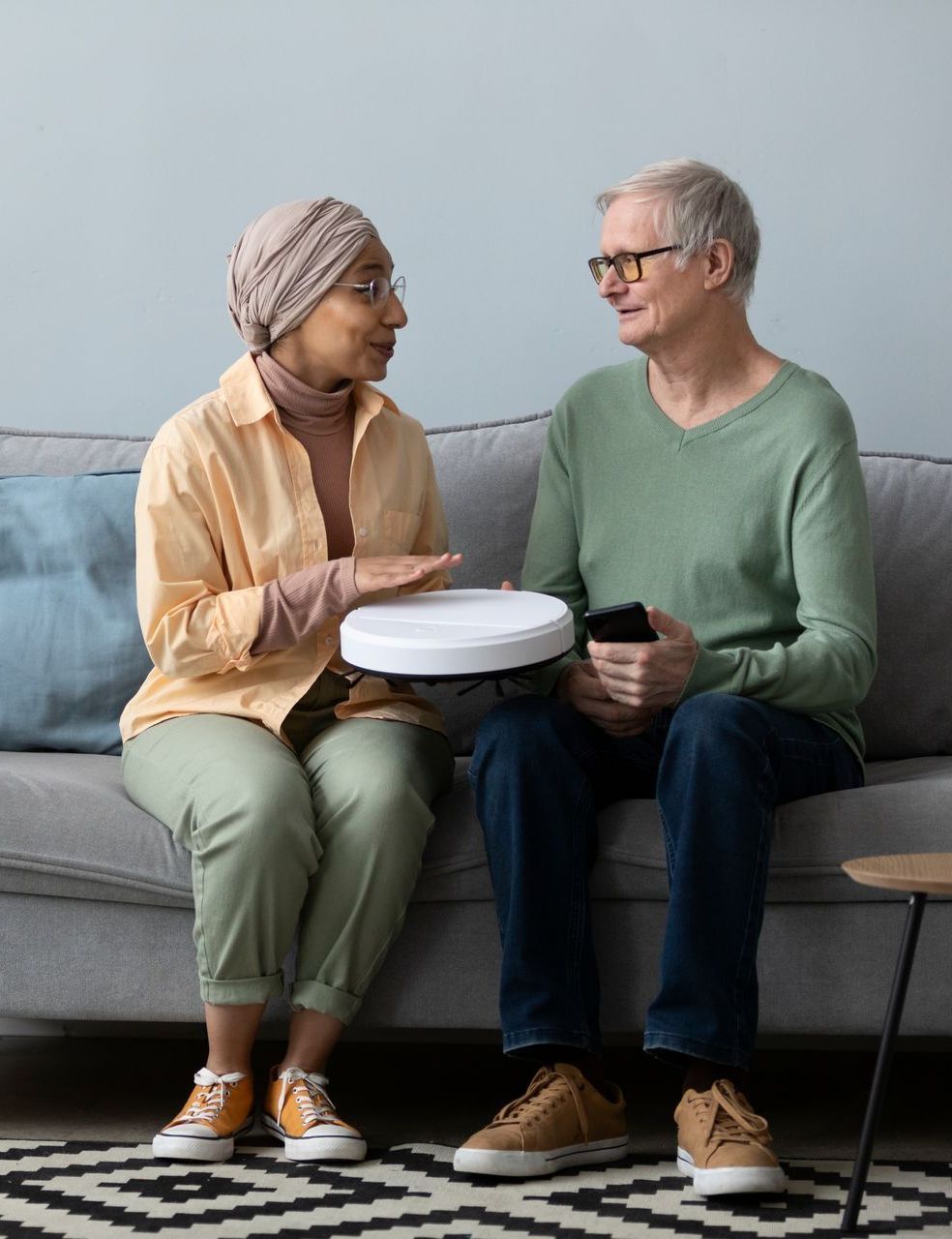 Two people sitting on a sofa, holding a robot vacuum. Woman in headscarf gestures, man holds phone.