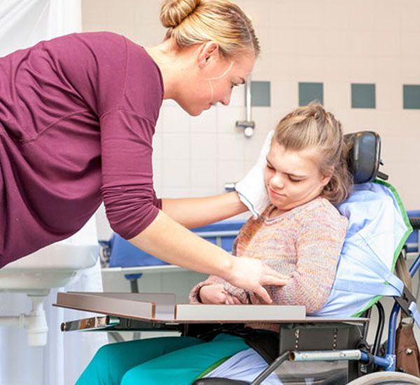 Woman assisting a young girl in a wheelchair, wiping her face with a washcloth. Bathroom setting.