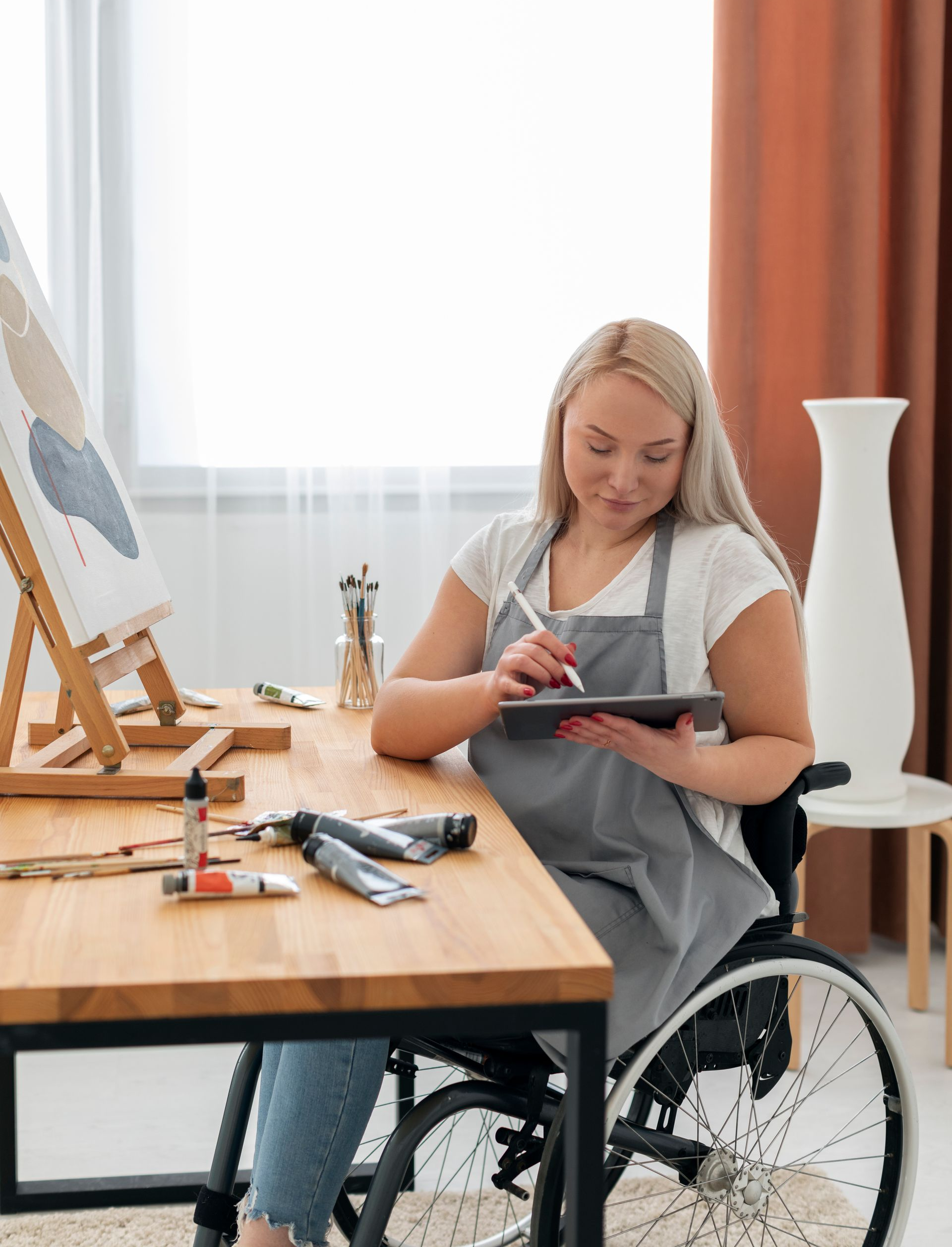 Woman helps an elderly woman from a wheelchair to a bed in a bright room.