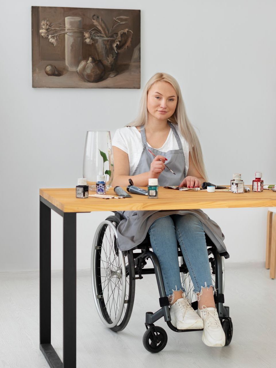 Woman with blonde hair, in wheelchair, painting at a wooden table. Gray apron, white shirt, jeans, artwork on wall.