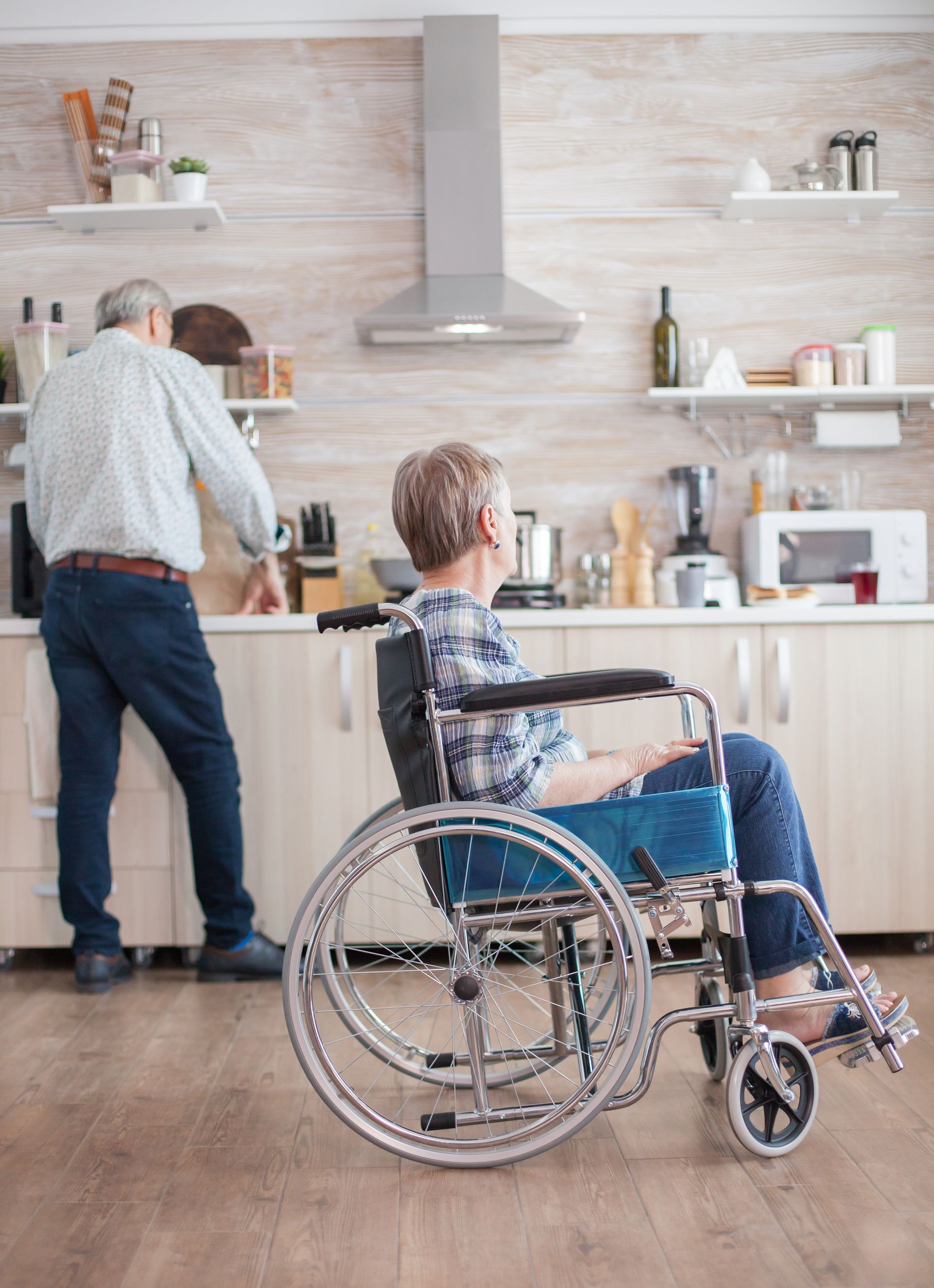 Elderly woman in wheelchair in kitchen, looking at man preparing food.