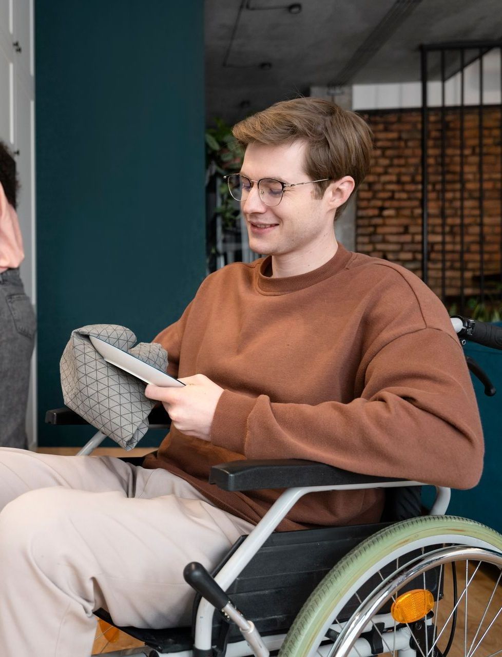 Man in wheelchair smiling at tablet, brown sweater, indoors.