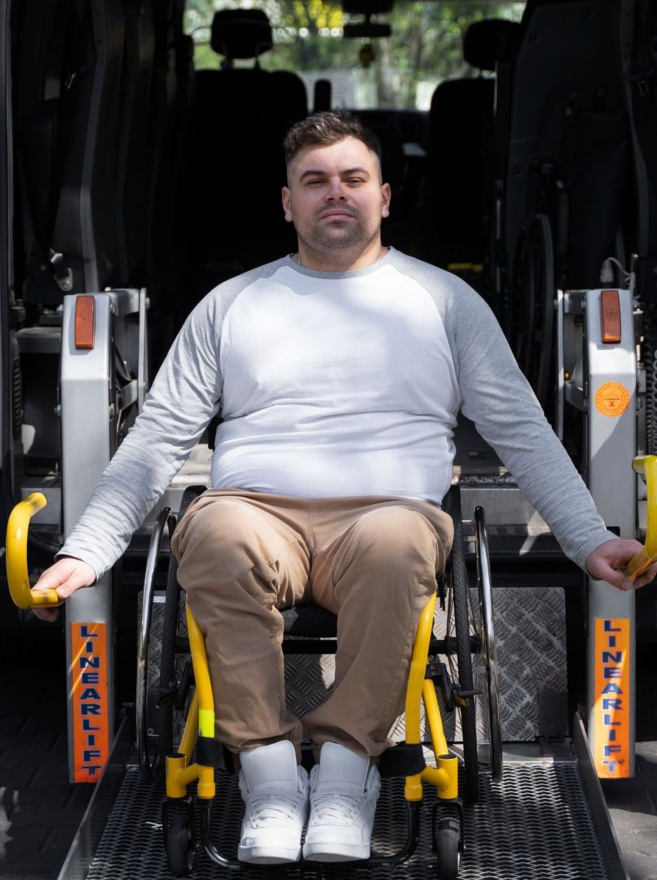 Man in wheelchair using a lift to enter a van. He wears a gray shirt, tan pants, and smiles.