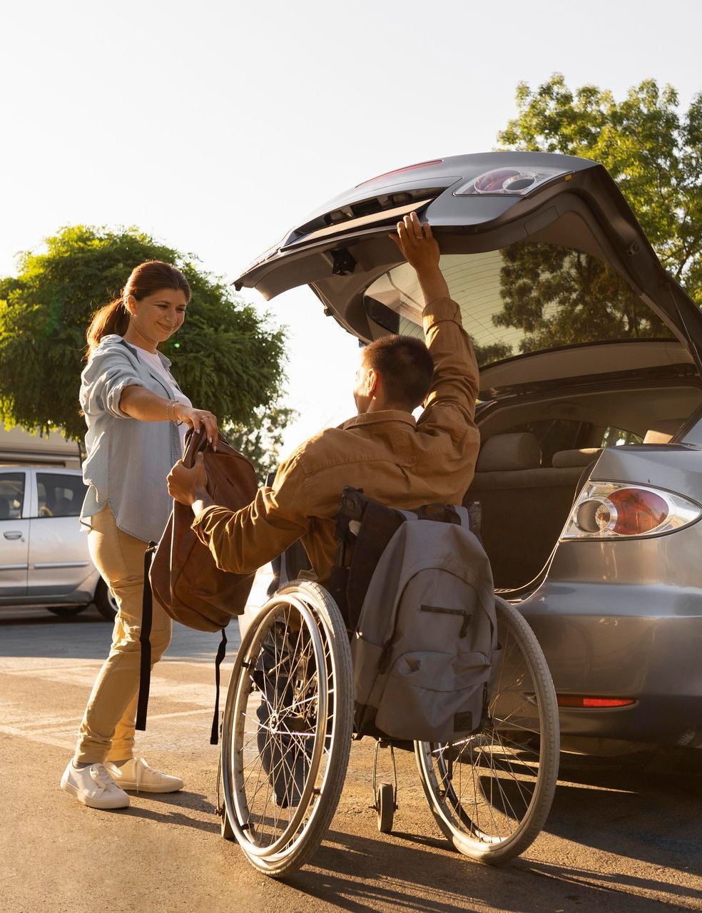 Woman helping a man in a wheelchair load a bag into a car trunk in daylight.