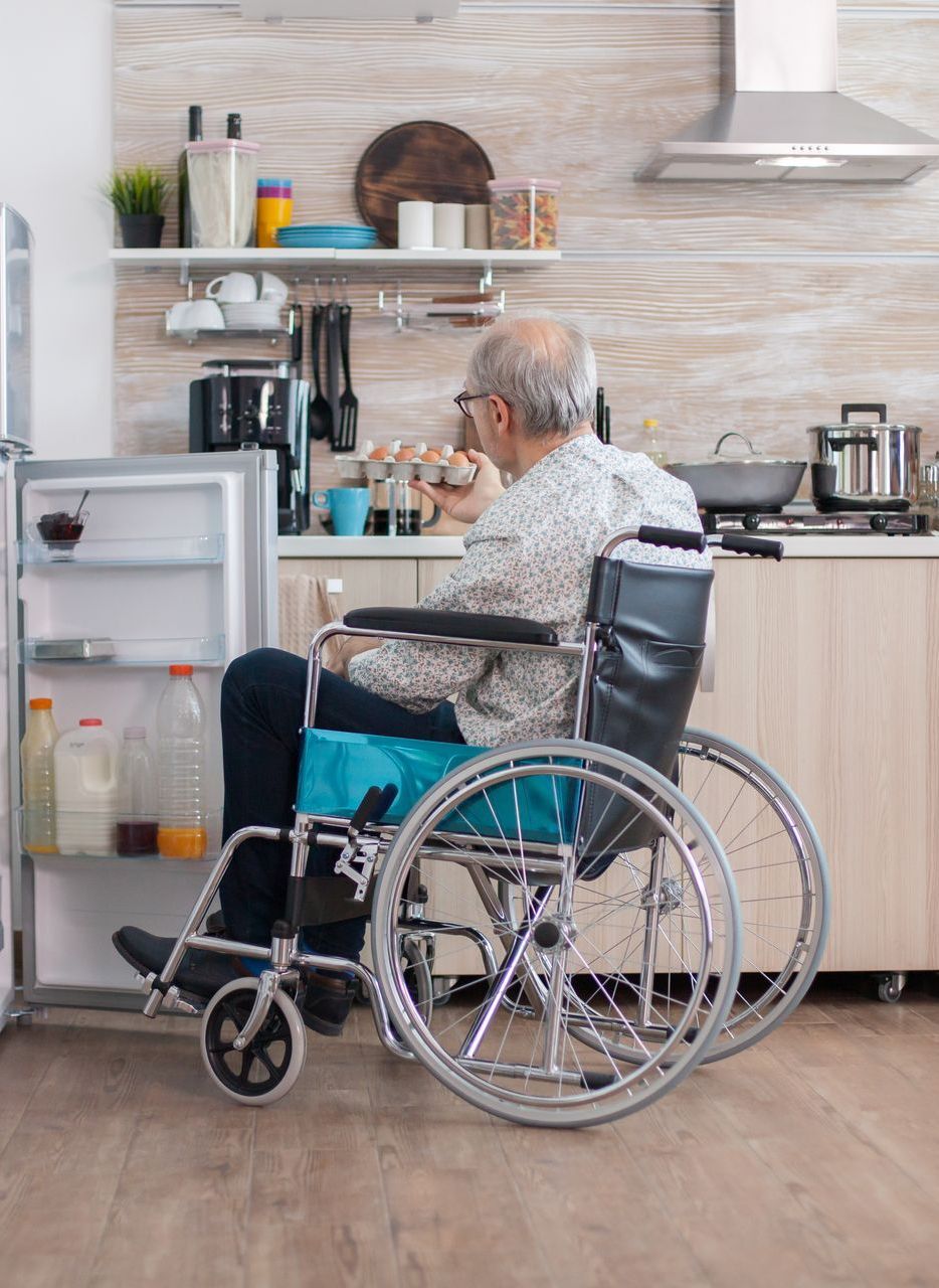 An older man in a wheelchair reaches into an open refrigerator in a kitchen.