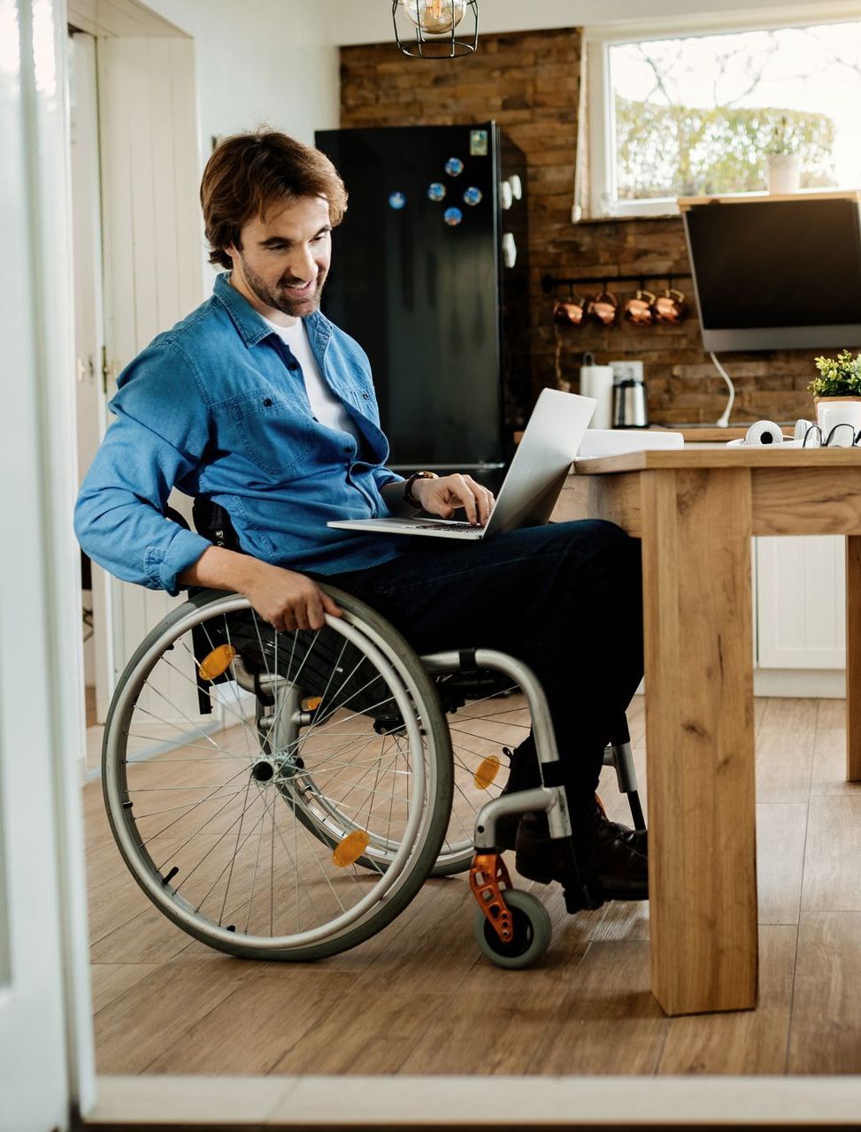 Man in a wheelchair using a laptop at a wooden table in a kitchen. He is smiling and wearing a blue shirt.