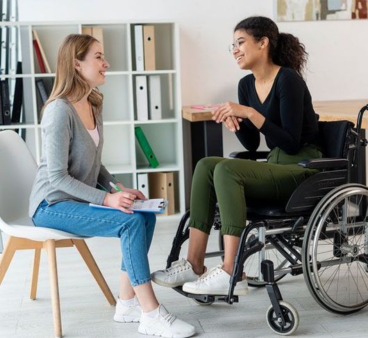 Woman in wheelchair talking with woman in office setting. Both smile.