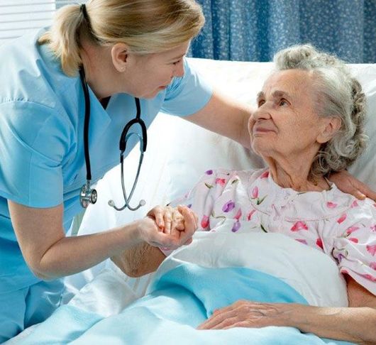 Nurse in blue scrubs holds hands with elderly patient in bed, offering support.