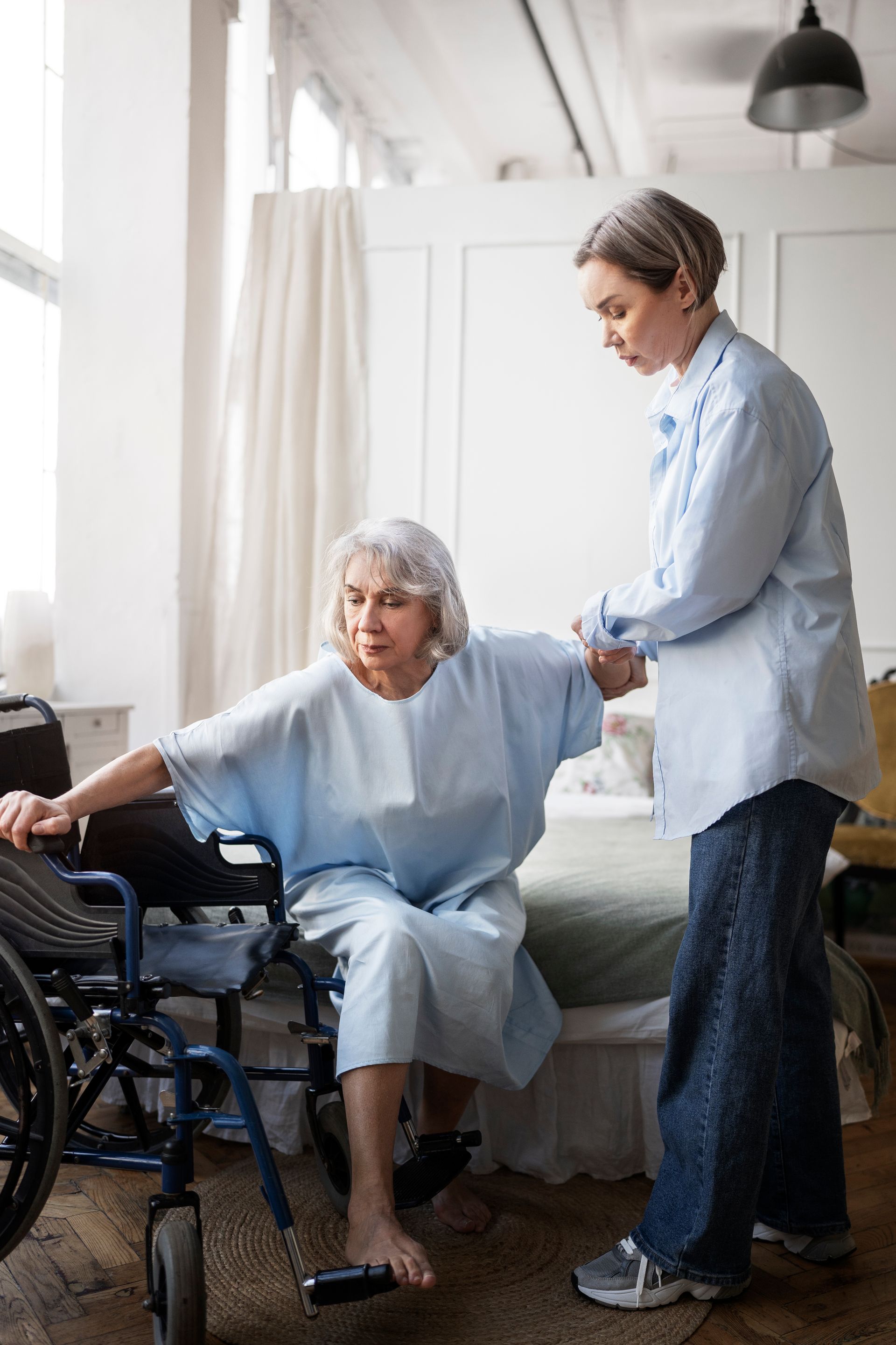 Woman helps an elderly woman from a wheelchair to a bed in a bright room.