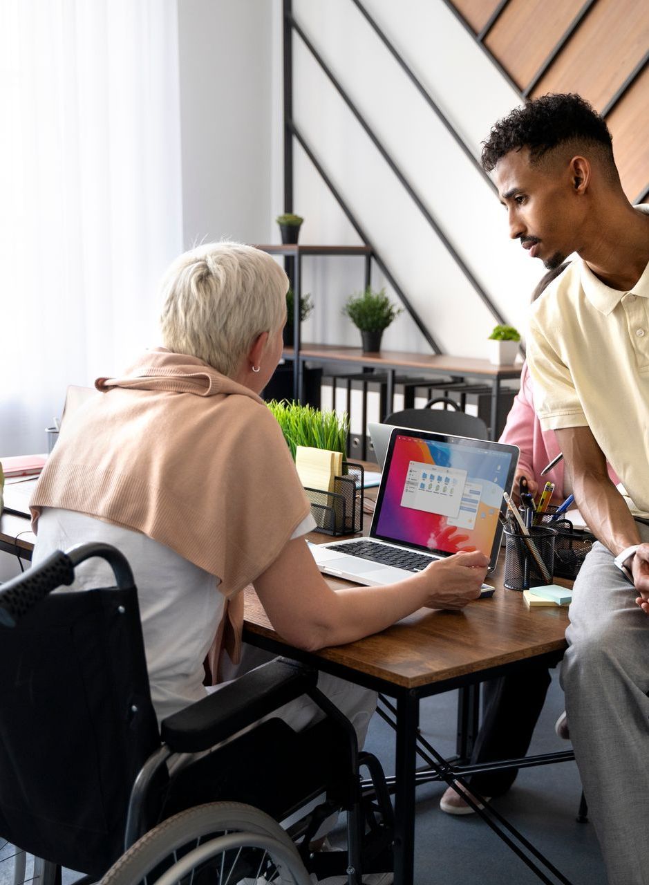 Woman in wheelchair points to laptop, discussing with a man in a bright office.