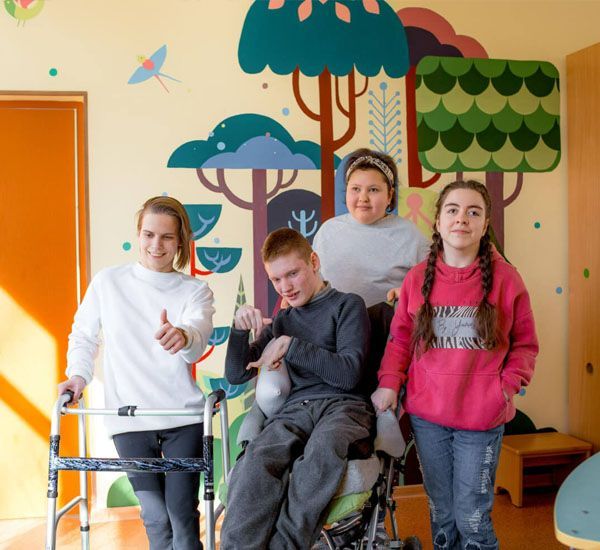 Four young people, some with disabilities, pose smiling in front of a colorful mural.
