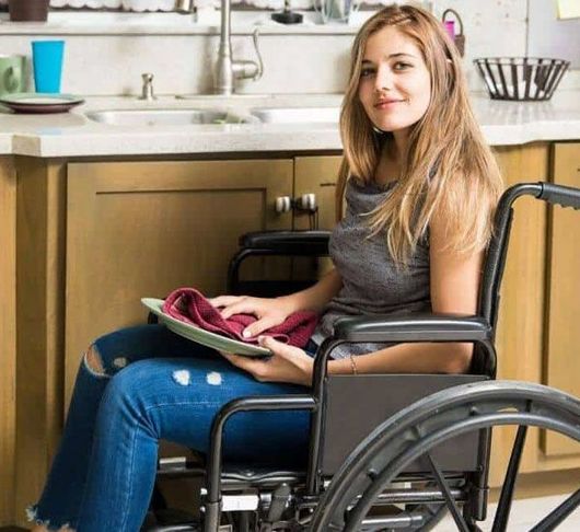 Woman in wheelchair washing dishes at a kitchen sink.