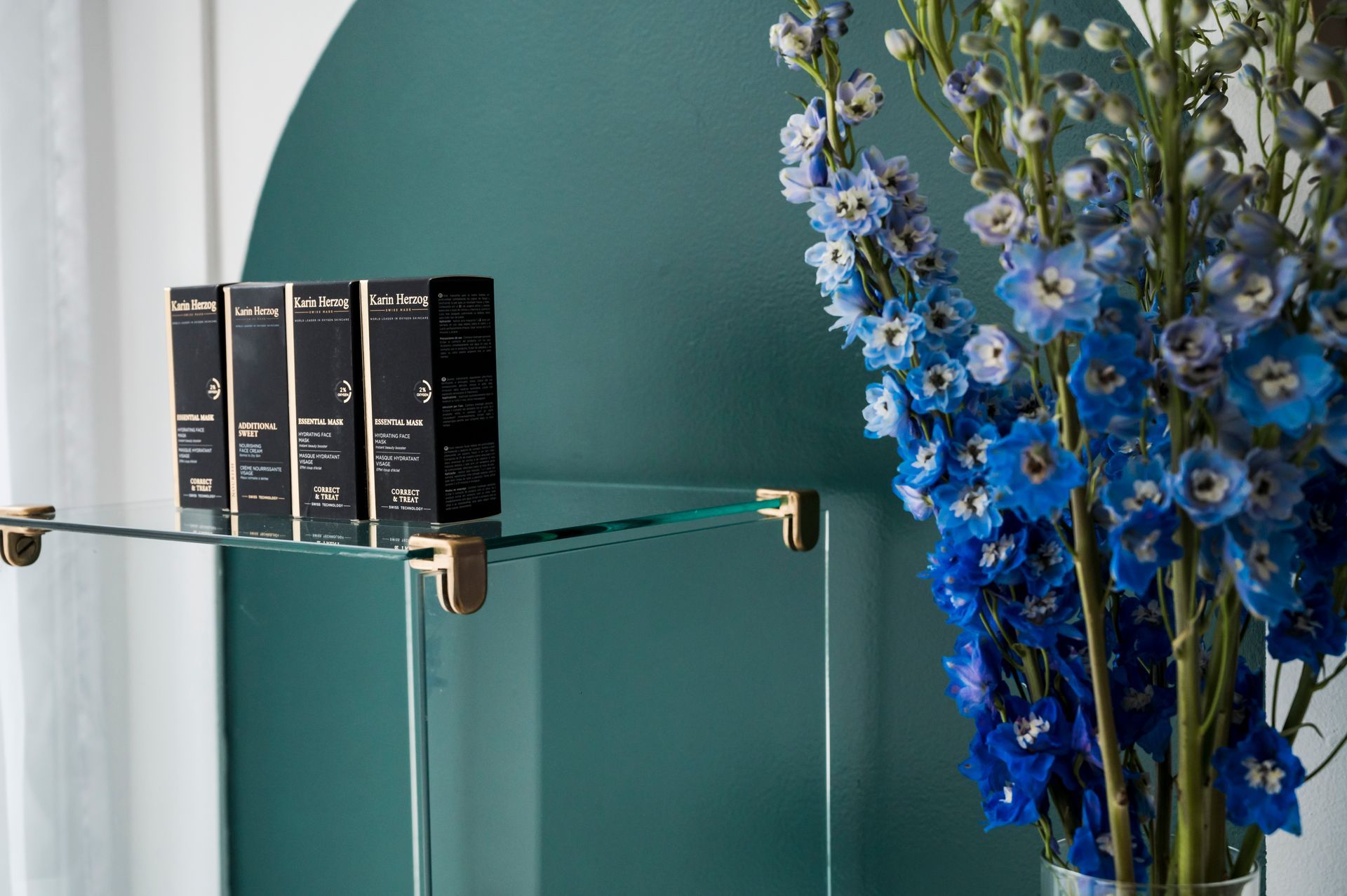 Glass shelf with beauty products and blue flowers against a teal wall.
