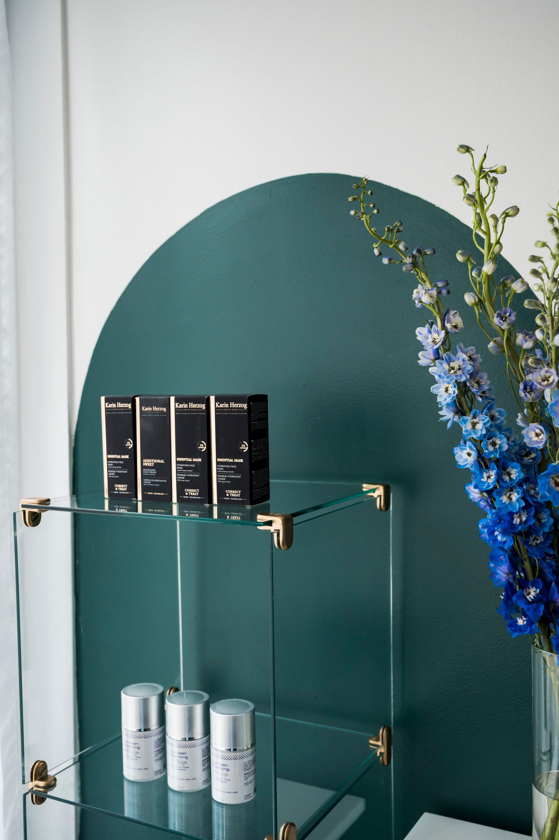 Glass shelves with skincare products against a teal arched wall, blue flowers in a vase.