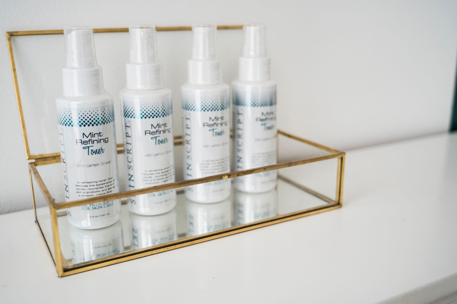 Four white spray bottles in a gold-framed glass box on a white surface.