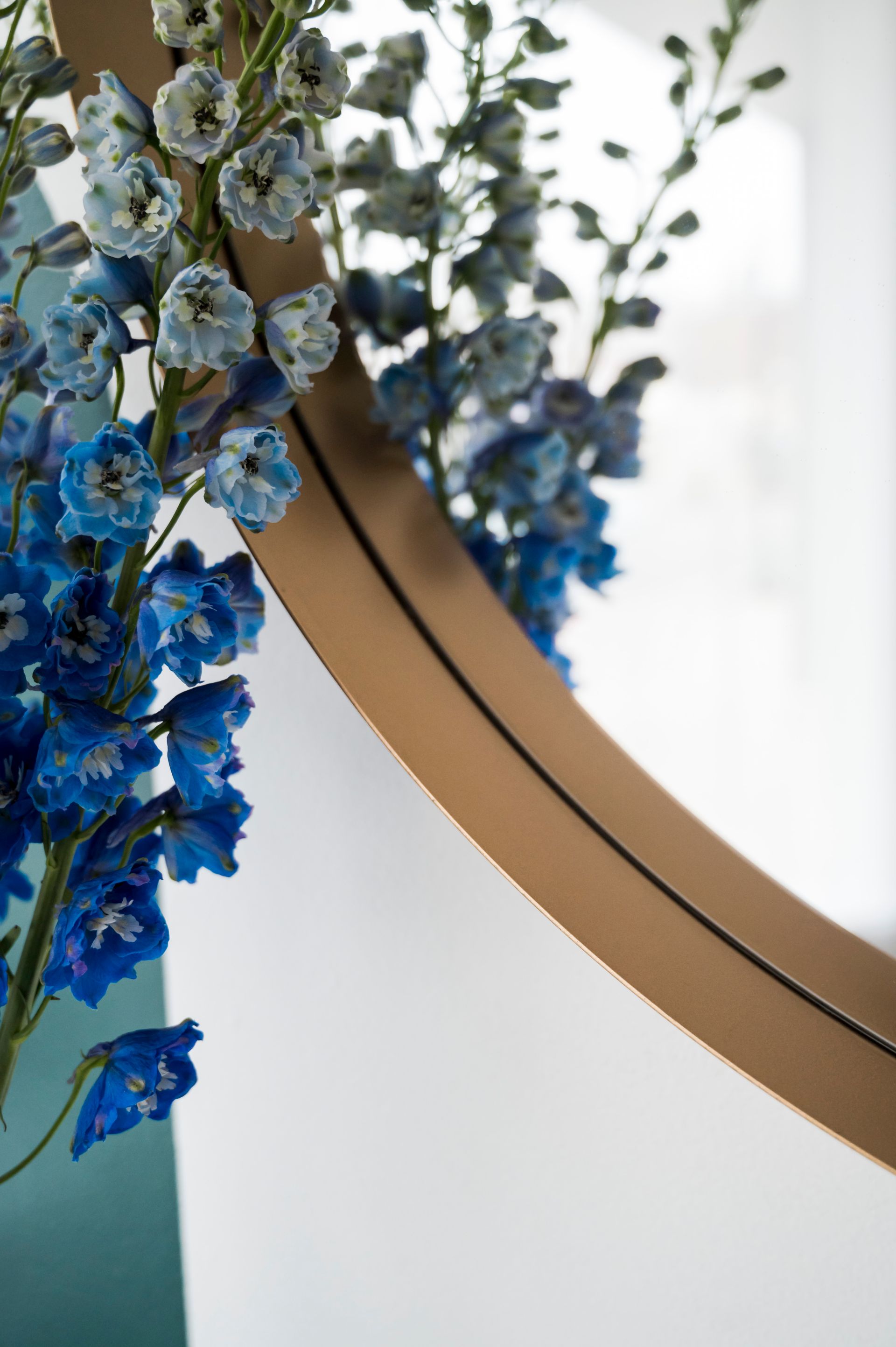 Blue flowers cascade near a round mirror with a gold frame, against a white and teal background.