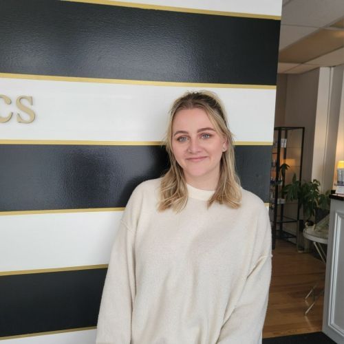 Woman in cream sweater smiles, standing near a black and white striped wall with gold trim.