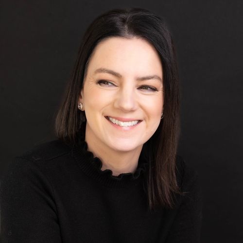 Woman with dark hair smiles, wearing a black top against a dark background.