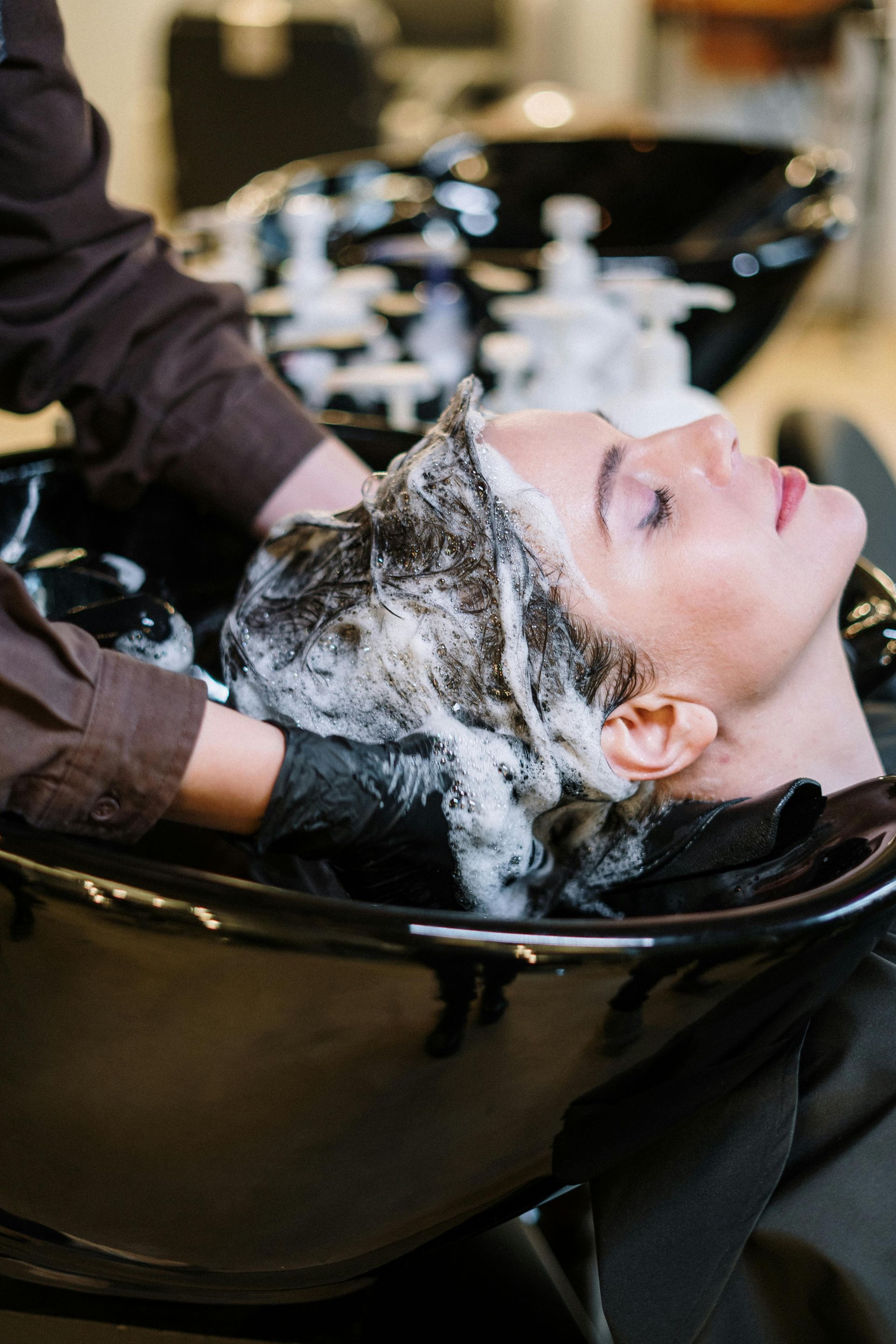 Person getting hair washed in a salon sink; dark hair with foam, gloved hands, relaxed expression.