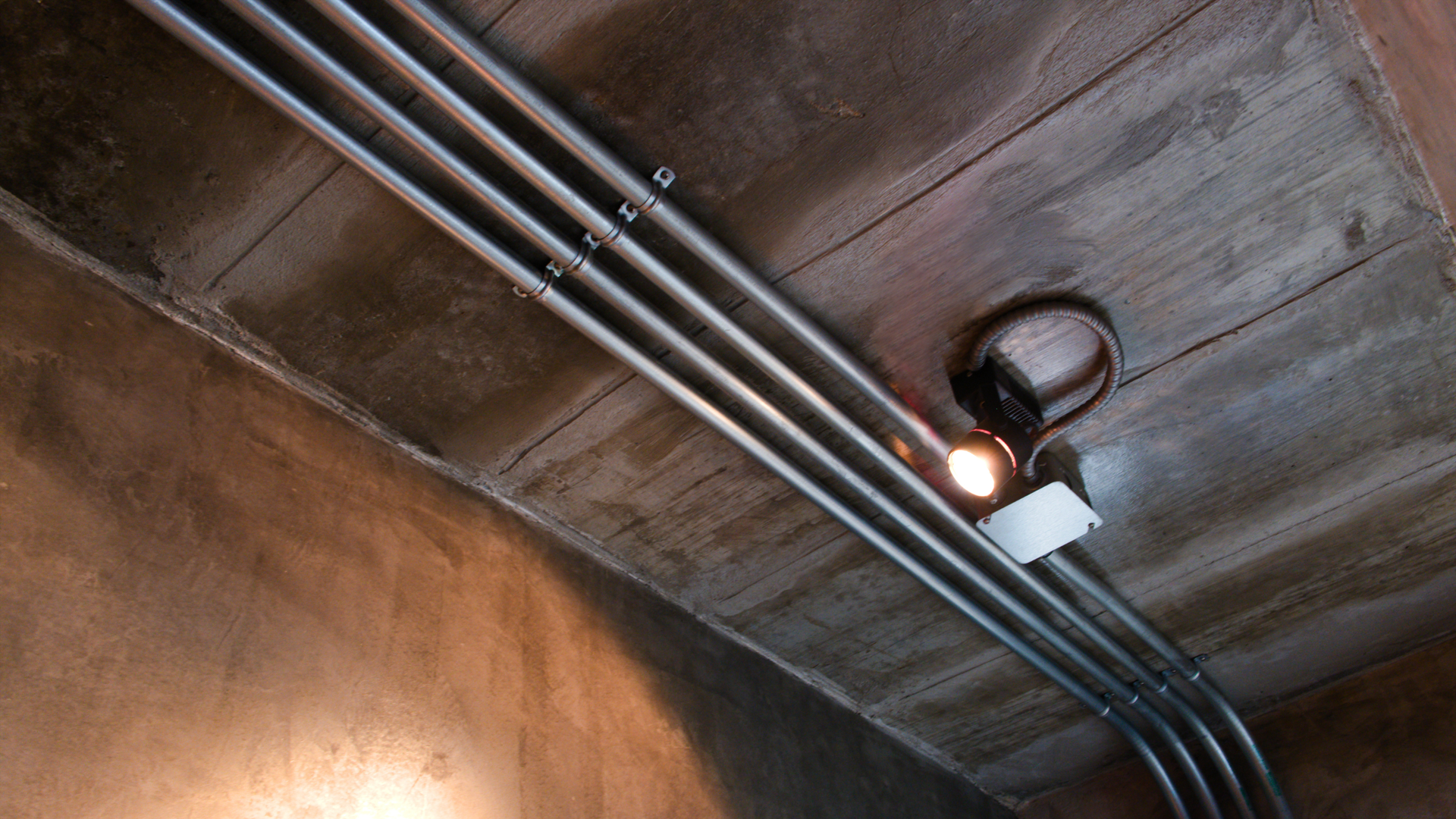 Three metal electrical conduits run along a textured ceiling, passing a small, lit spotlight attached to a circular base.
