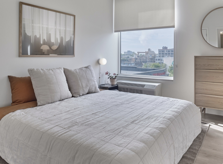 Bedroom with bed, art, window, dresser, and round mirror. Neutral tones, natural light.
