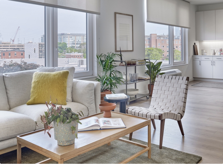 Living room with white sofa, wood coffee table, woven chair, plants, and large windows.