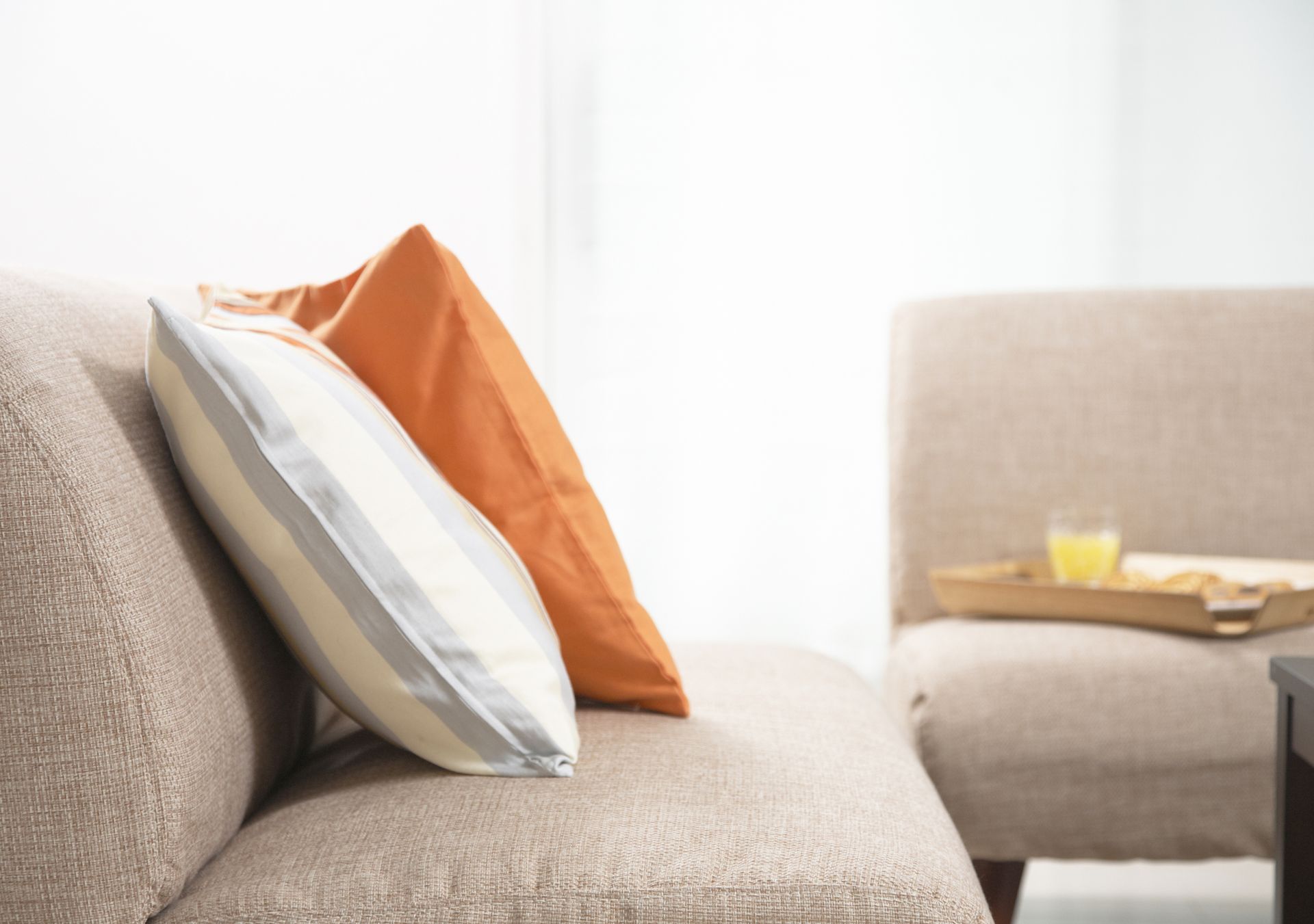 Beige sofa with orange and striped pillows, chair, tray with drink.