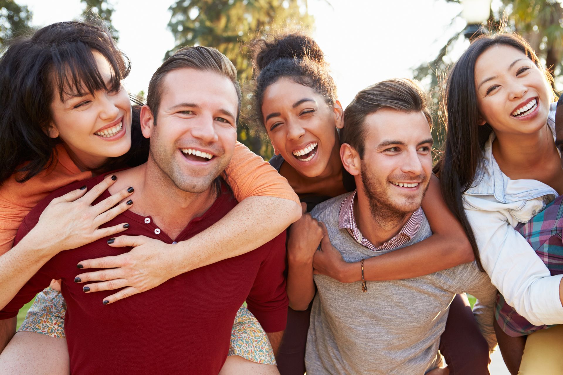 Group of smiling people, two giving piggyback rides outdoors, sunny background.