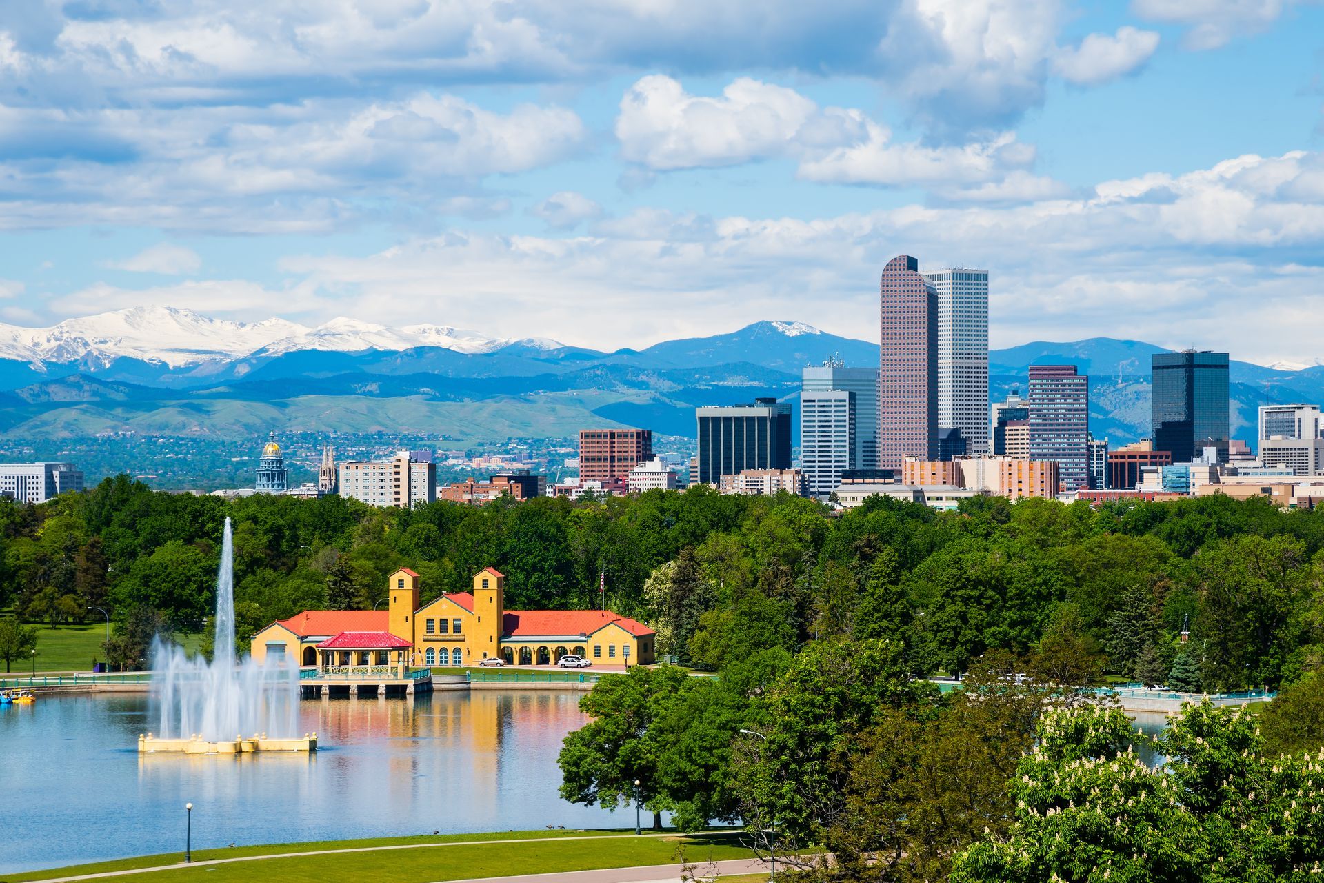 Denver skyline over a lake and park, with mountains in the background under a blue sky.