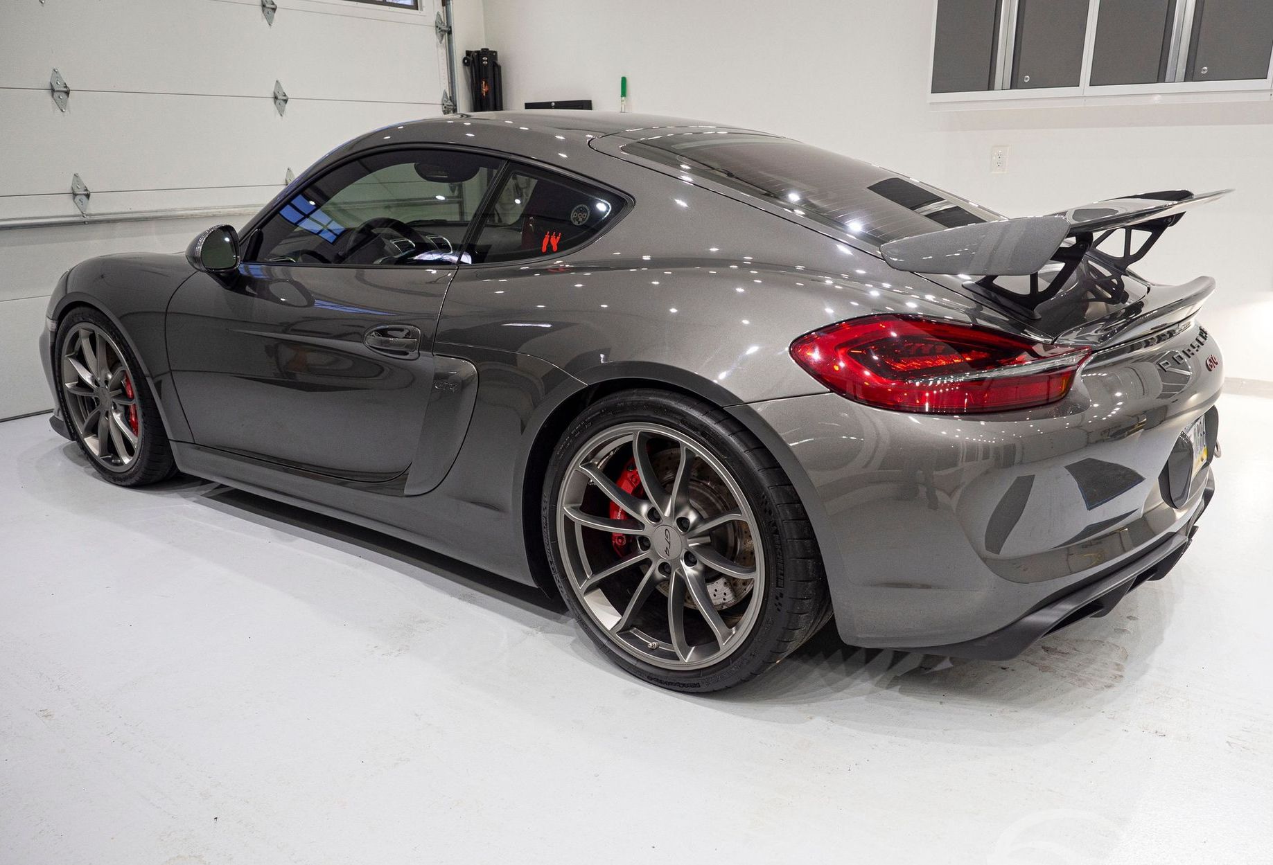 Gray Porsche Cayman with a rear spoiler parked in a garage with a white floor.