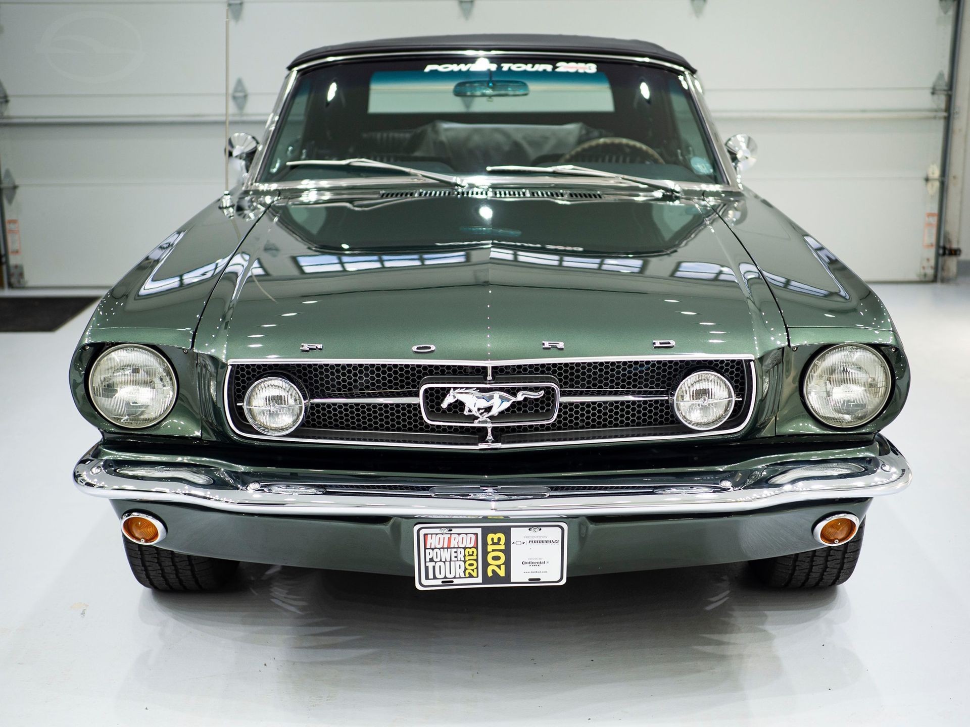 Green vintage Ford Mustang convertible, front view, parked indoors. Chrome details and a black top.