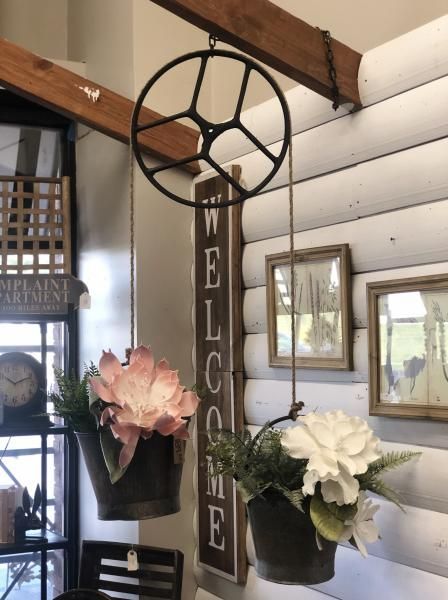 Rustic porch wall with vertical “WELCOME” sign and hanging flower baskets beneath wooden beams