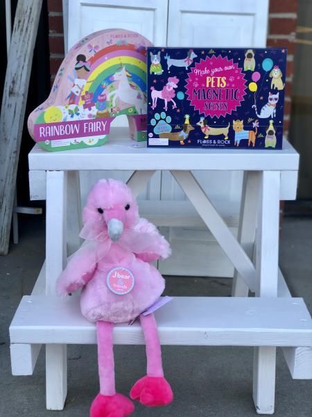 Pink stuffed flamingo seated on white steps beside two colorful toy boxes and a rainbow-themed bag