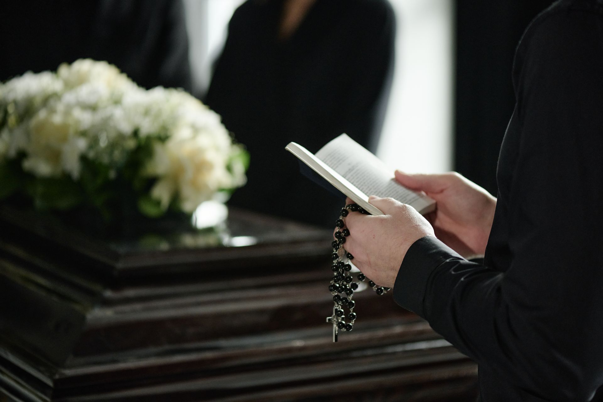 Person holding a prayer book and rosary beside a casket with white flowers at a funeral