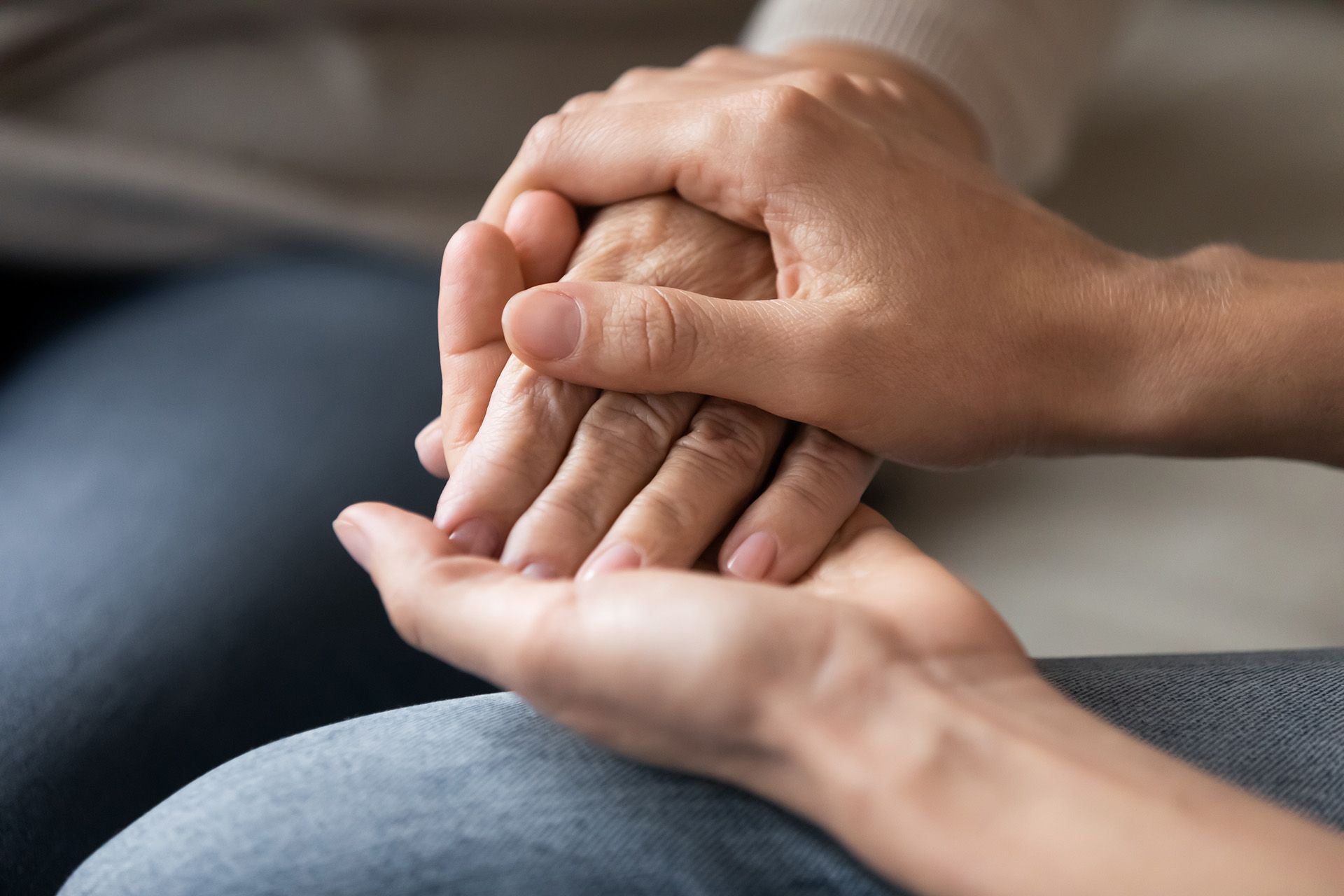 Two hands clasped gently over another hand on a lap, suggesting support or comfort