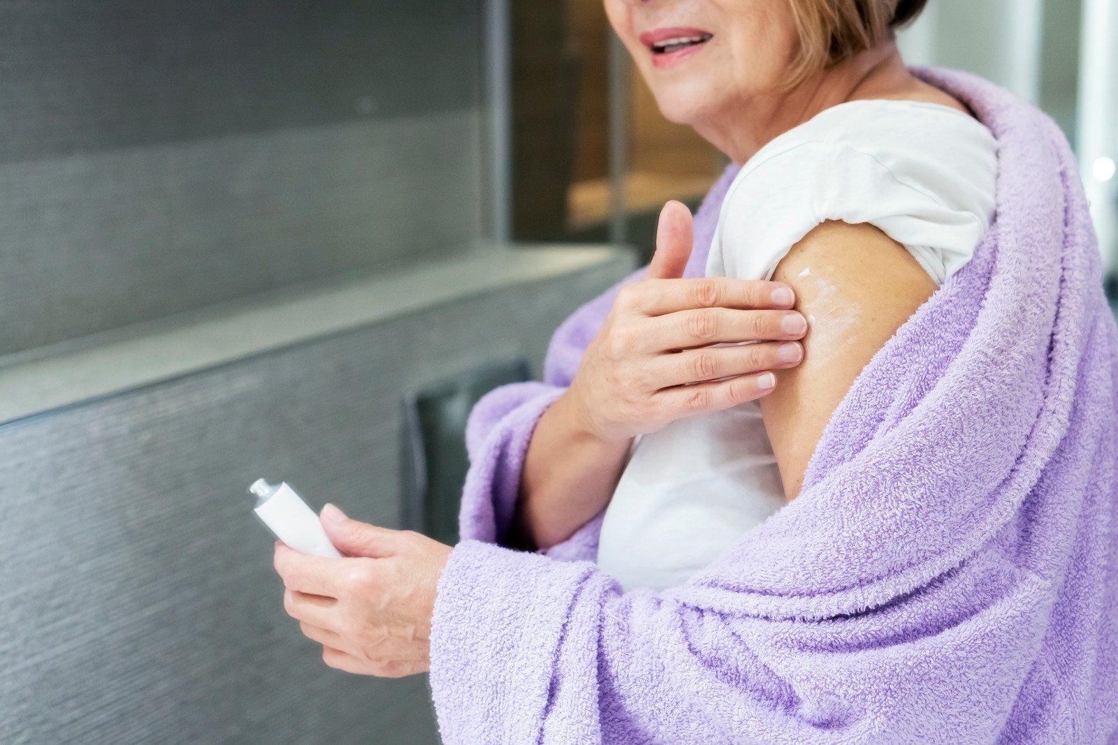 A woman wrapped in a purple towel is applying lotion to her arm.