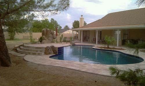 Backyard pool with rock waterfall feature, steps, and house with a brown roof.