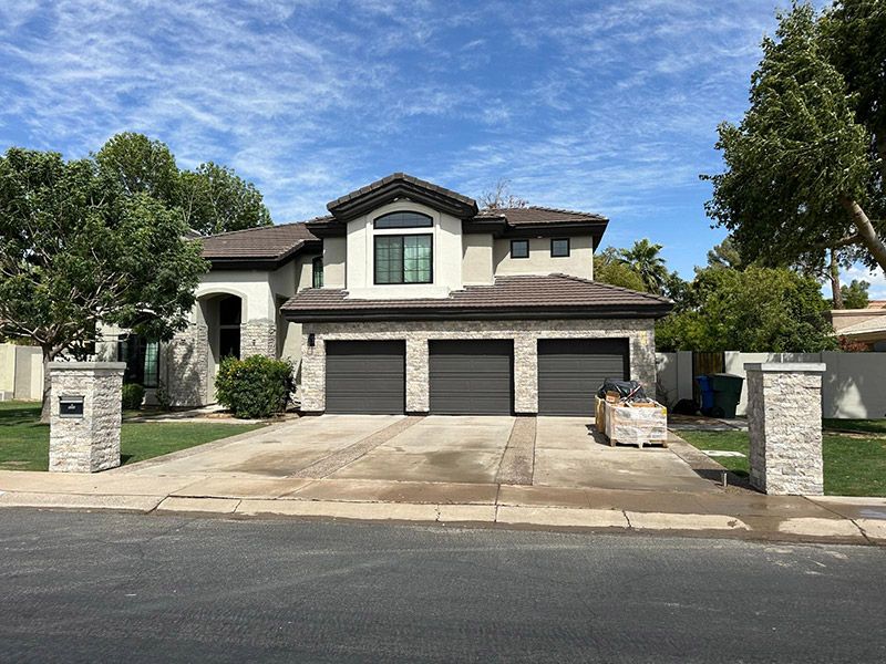 Two-story stucco house with three-car garage. Driveway, green lawn, trees, blue sky. Stone columns at entrance.